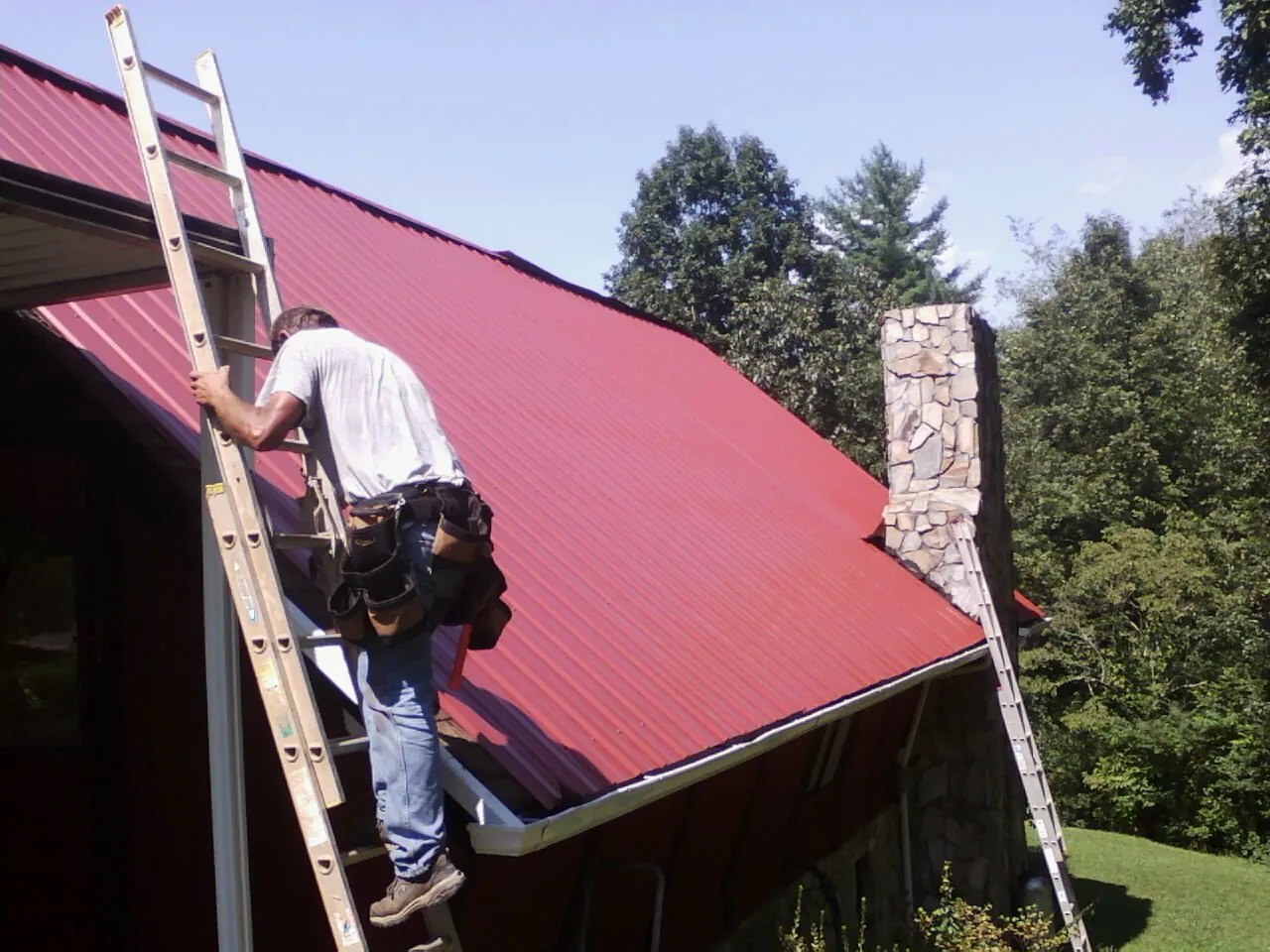 Roofer on a ladder beside a red metal roof and stone chimney — Soffit & Fascia Repair by Roofing Brothers in Hildebran